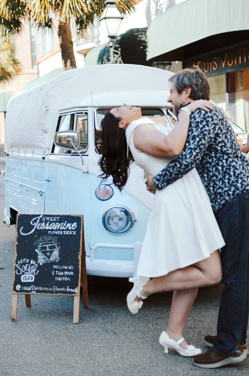 A bride and groom are laughing and holding each other in front of a blue flower truck.