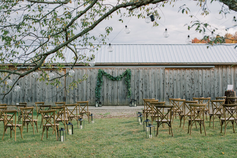 Rustic outdoor wedding ceremony setup with wooden cross-back chairs, lantern aisle decor, and greenery arch in front of a barn at Bear Ridge Destination.

