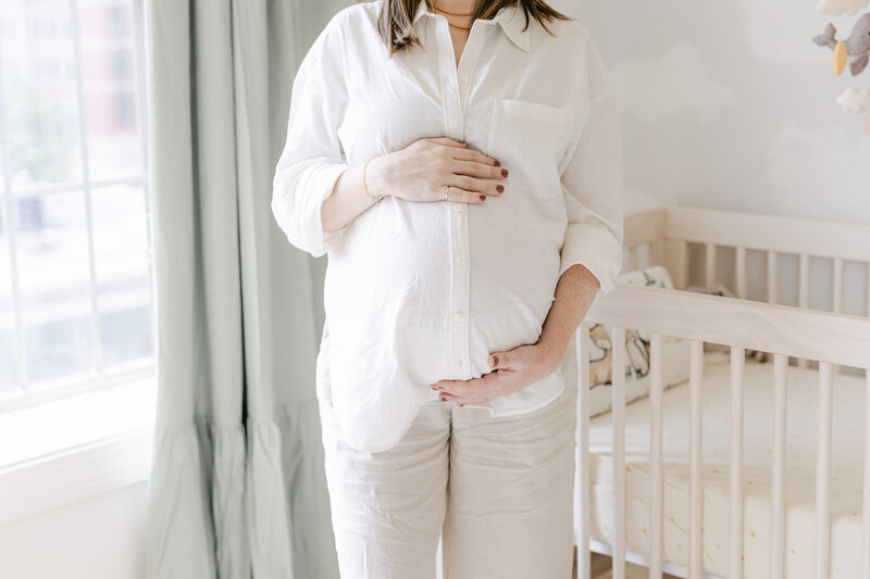 A pregnant mother cradles her belly while standing in a softly lit nursery, wearing a white button-down shirt and linen pants. Captured by Nicole Neira Photography in Fairfield County, CT, this in-home maternity session highlights the quiet anticipation before baby’s arrival.