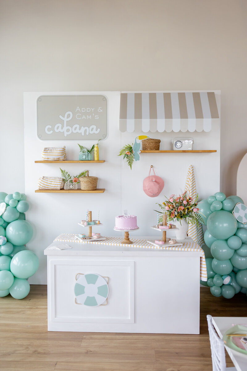 Beach wall with striped cabana, name sign, beach flowers, pink desserts, cakesicles and blue balloons and lifesavers.