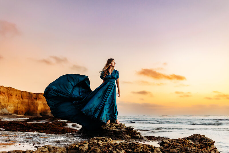 High school senior girl in a billowy dress looking out to sea at sunset.