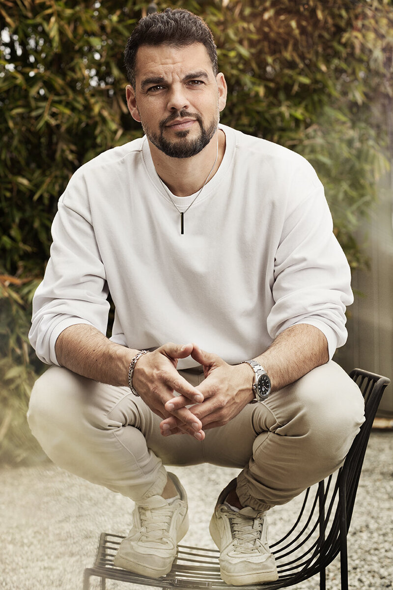 Casual outdoor portrait of David Victori in a relaxed pose, wearing a white sweater and beige pants, captured by Ylva Erevall.