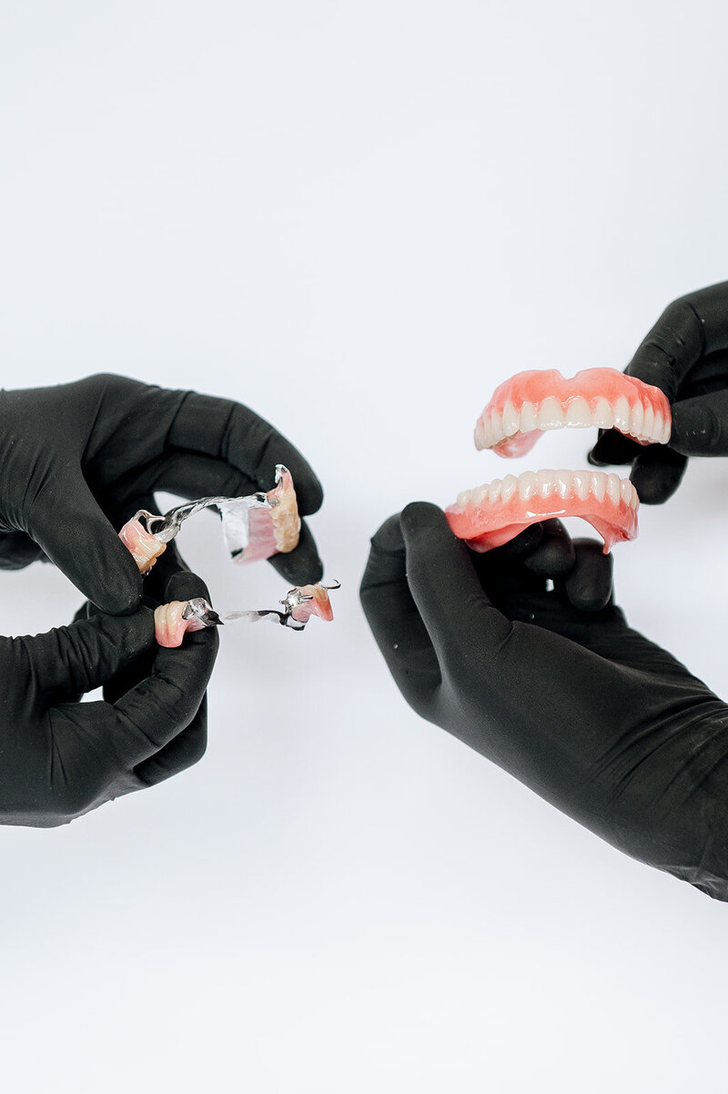 Four hands wearing black gloves hold partial and full dentures.
