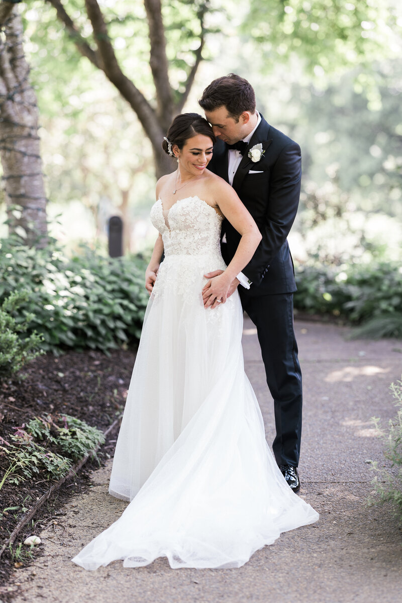 a white groom in a black tux holding a white brunette bride in a strapless white gown nuzzling her neck and her smiling