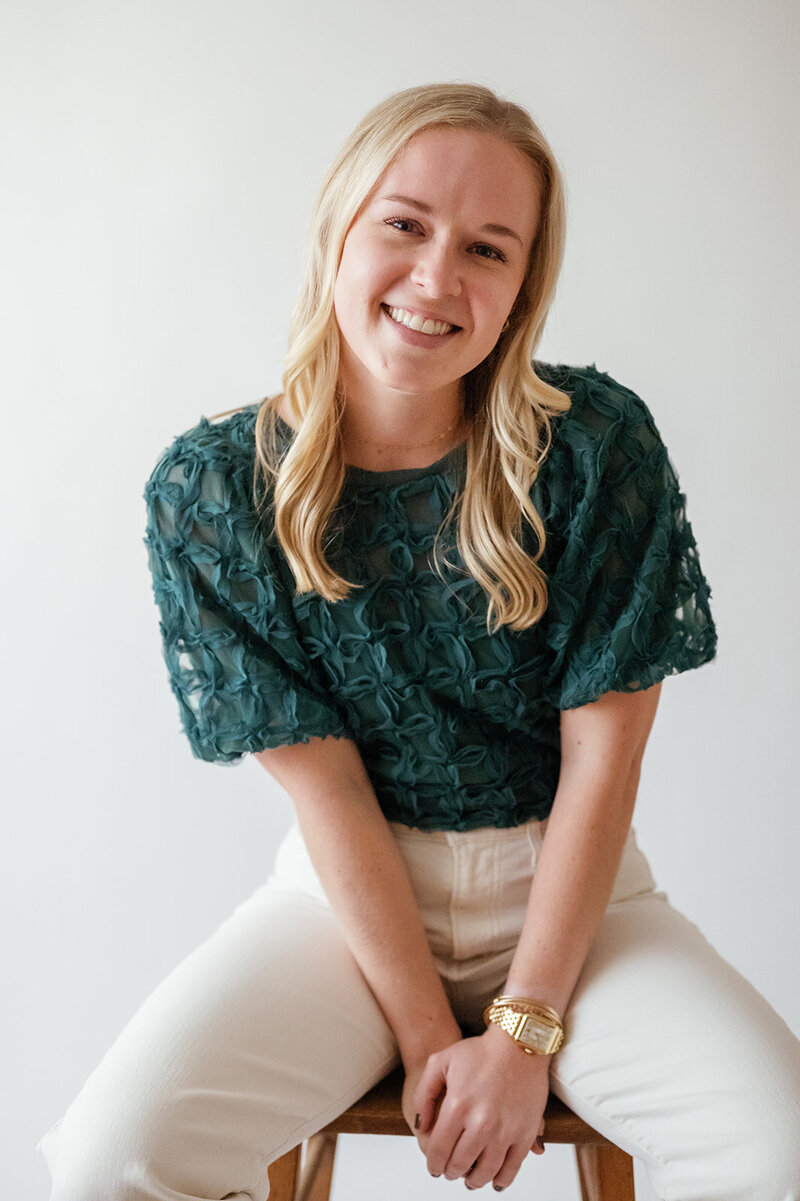 Woman in a teal top and white pants sitting on a stool