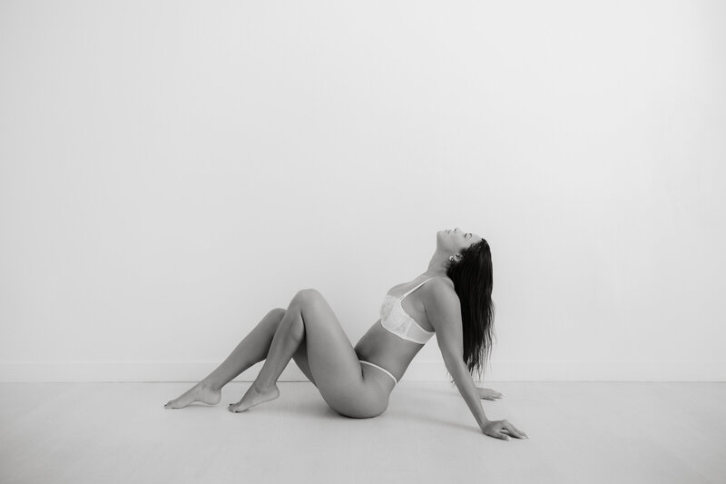 Black and white photo of woman in lingerie sitting on the floor leaning back in front of a white wall