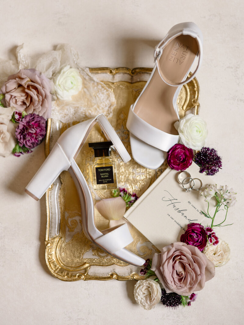 White open-toe heels on a gold tray, surrounded by roses and a small bouquet. A bottle of perfume and a handwritten note with elegant script accompany the arrangement, creating a romantic, elegant setting.