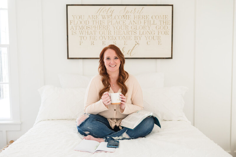 A faith-led business coach sitting cross-legged on a bed with scripture artwork in the background, representing Ashleigh Foy — a systems and efficiency coach helping Christian women build sustainable, faith-aligned businesses rooted in purpose and peace.