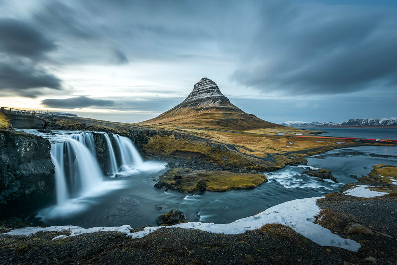 Scenic view of a pointed mountain with a waterfall and river in the foreground under a cloudy sky.