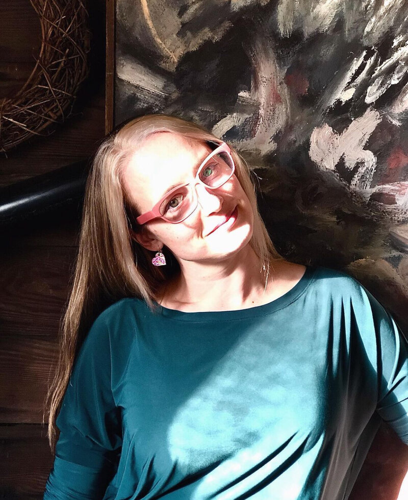 woman in teal shirt and pink glasses smiling in front of a painting