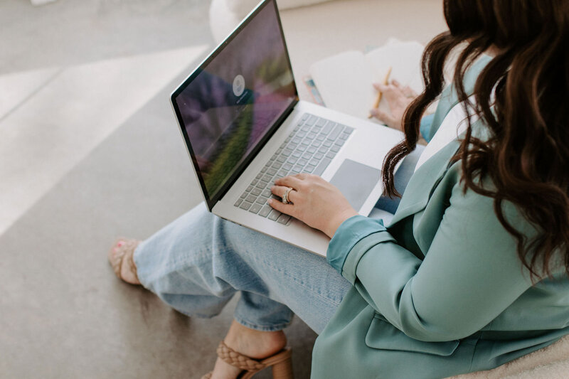 Woman with dark hair, light teal blazer and jeans working on her laptop