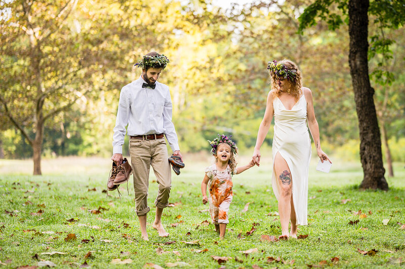 A family of three walk in a park together to head to  their ceremony spot for their elopement day. One parent holds the hand of her child while the other parents  holds two pairs of shoes in his hands.