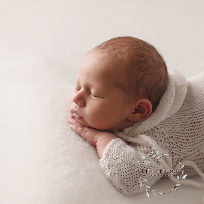 profile photo of a newborn boy dressed in a white knitted romper posed on his tummy with his hand under his chin