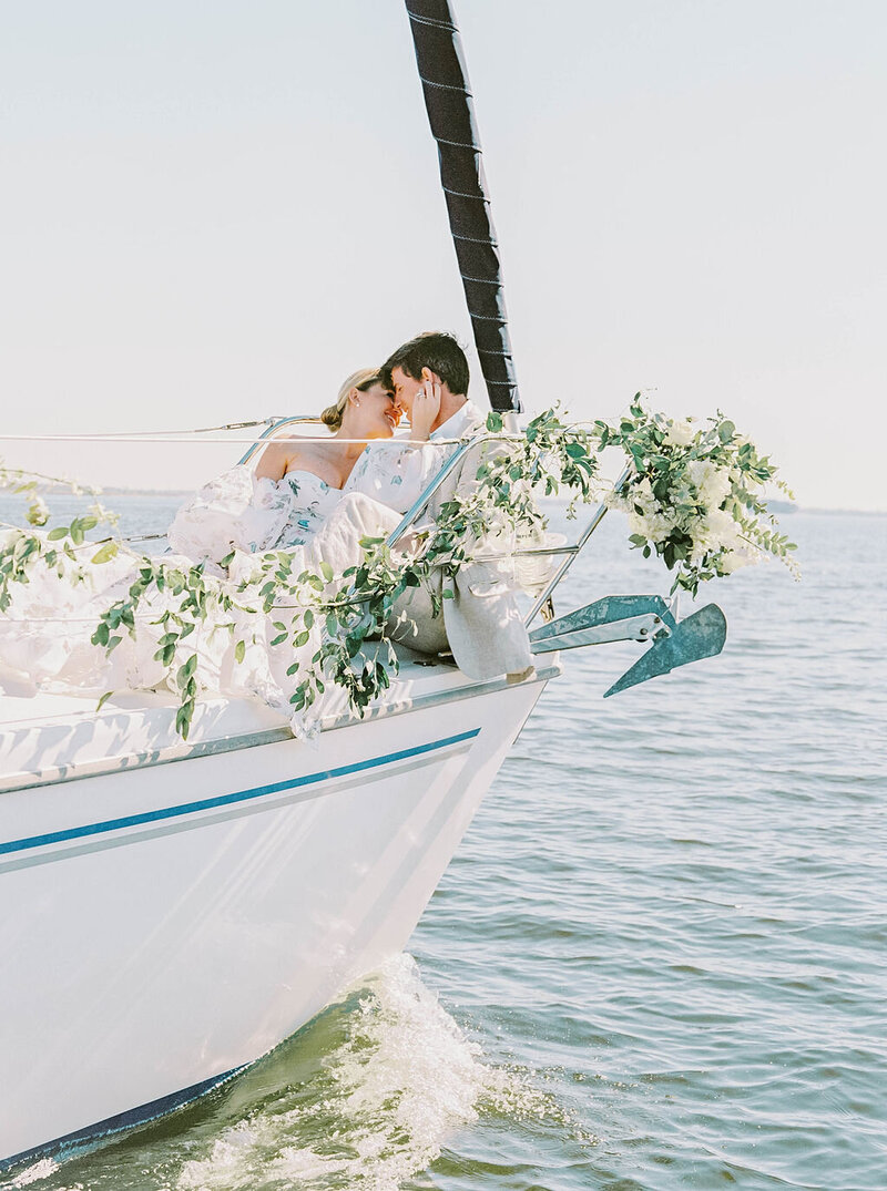 Newport RI Wedding Photographer | A couple shares a tender kiss on the bow of a sailboat adorned with lush white flowers and greenery, set against a serene blue ocean backdrop.