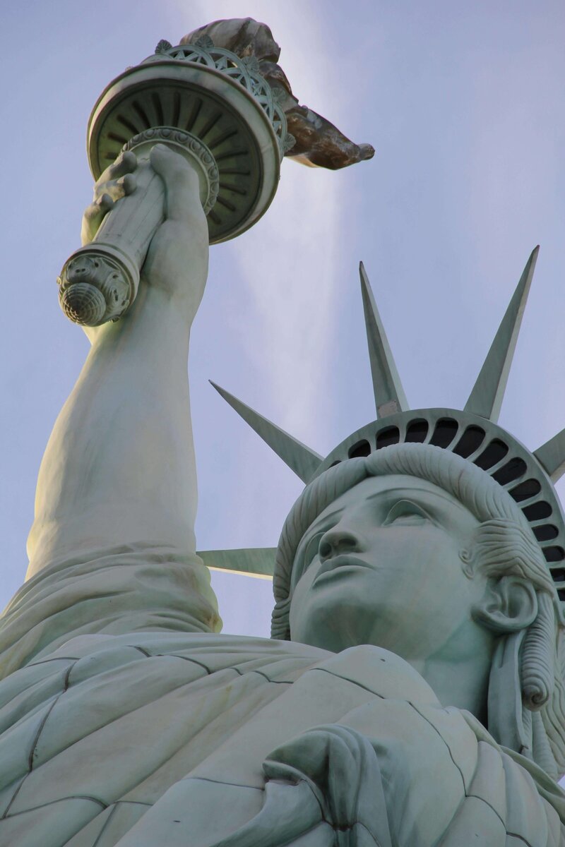 A striking upward view of the Statue of Liberty against a soft blue sky, emphasizing its iconic torch and regal expression. This symbol of freedom and hope highlights the cultural experiences that can be enjoyed through vacation fundraising for charities.