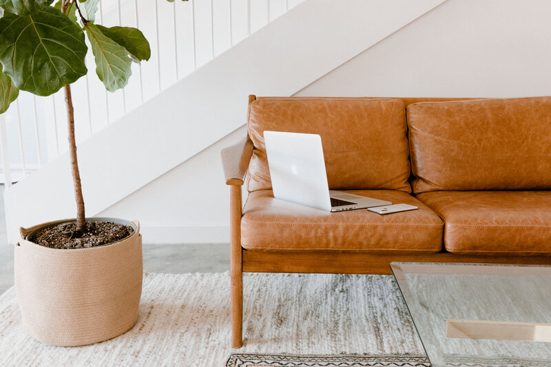 A tan leather sofa with a laptop and smartphone—perfect for online learning—sits by a large potted plant. The space inspires creativity, making it ideal to take a white label design course beneath the crisp white staircase and glass coffee table.