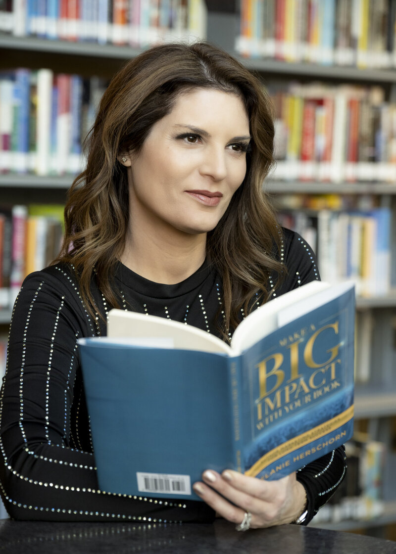 Melanie is in a black dress with rhinestones, seated in a library on holding a book open