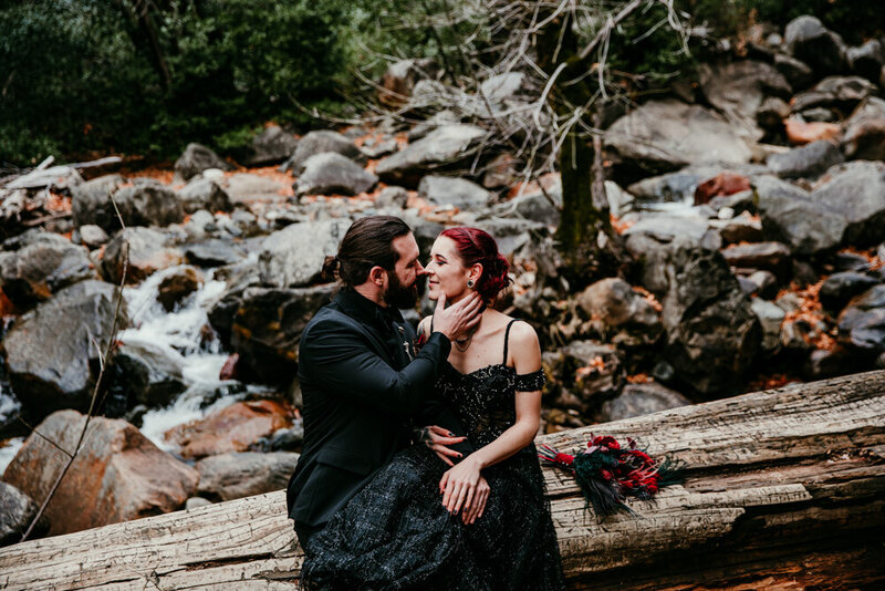 Couple in dark, elegant attire sharing an intimate moment on a fallen log near Bridalveil Fall in Yosemite National Park, with a rocky creek flowing in the background.