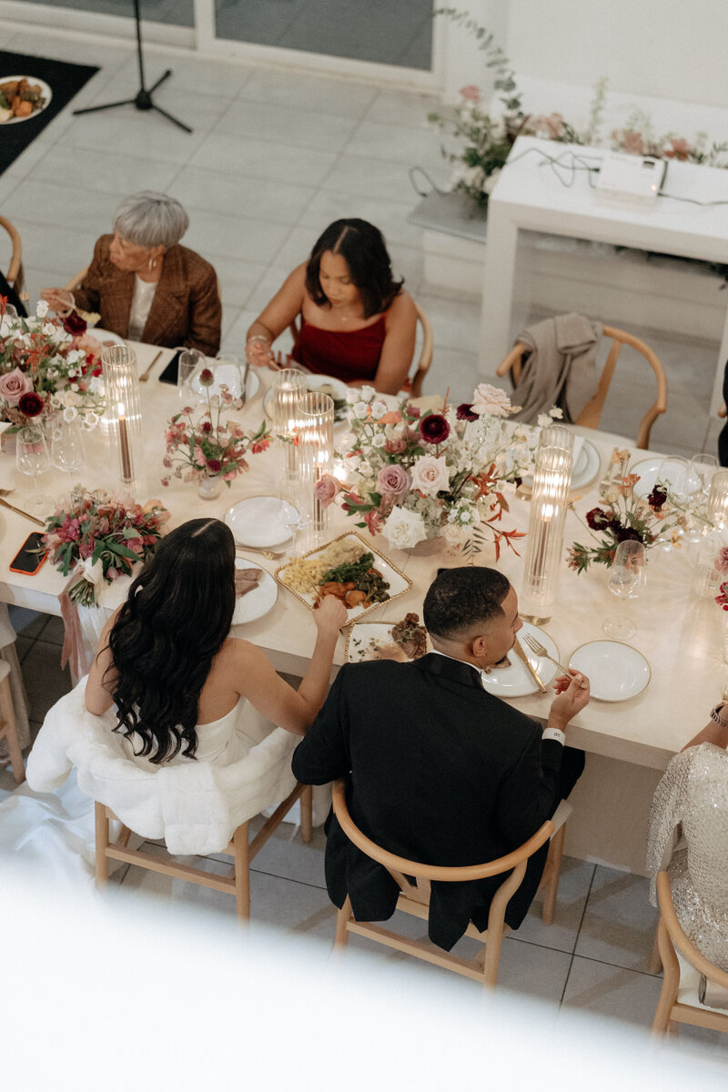 Overhead view of a wedding reception table with sculptural florals, ribbed glass candles, and guests dining on wishbone chairs in a Houston venue.