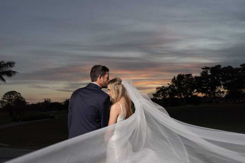 Bride and groom embrace with a flowing veil against a Miami sunset backdrop.