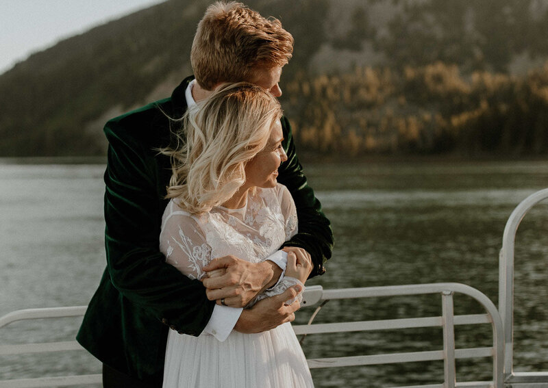 Travel photographer in Eastern Idaho, Adriana Montoya Photo. A groom is standing behind his bride and holding her from behind. She is holding his arms and they are both looking off to the right, so we can't really see their faces. Their standing on a bridge, with water behind them and in the far distance, trees on a mountain.