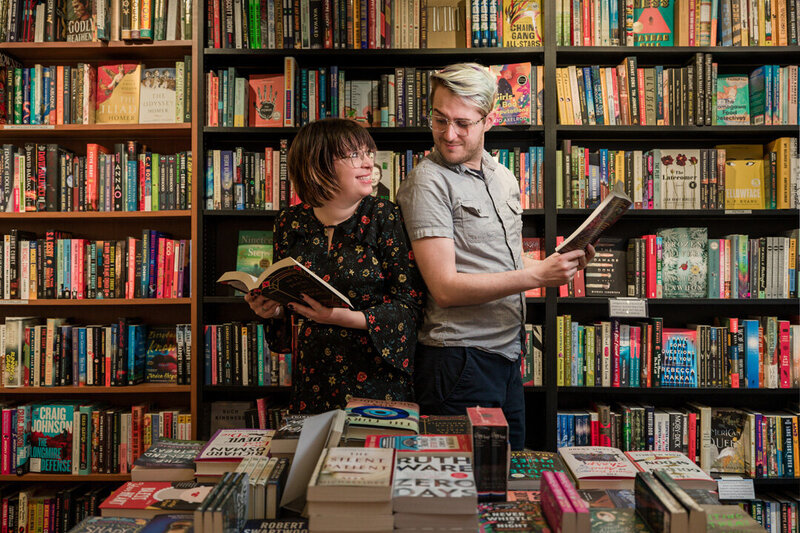 Couple sneaking peeks at each other in a cozy bookstore in Phoenixville, a playful and romantic engagement moment.