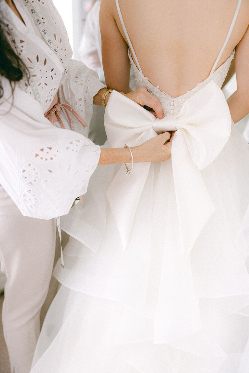 Bridal seamstress tying a large satin bow on the back of a layered tulle wedding gown during a dress fitting.