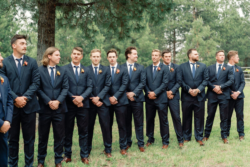 Groomsmen in navy suits wearing coordinating boutonnieres with coral-toned blooms and greenery at Kelly and Joe’s wedding, designed by Summer Robbins Flowers in Bend, Oregon”