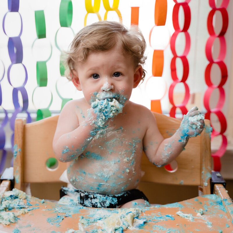 A toddler covered in icing sitting in a highchair while eating cake.