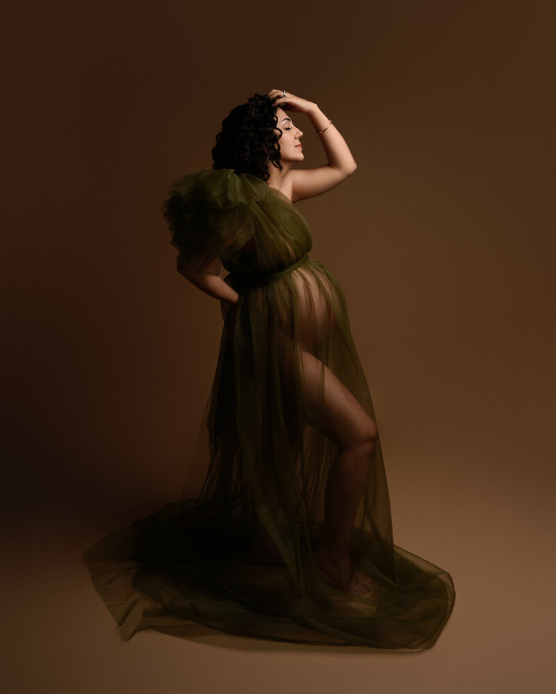 Textured olive green maternity dress with sheer layers and off-shoulder draping – photographed against warm brown backdrop