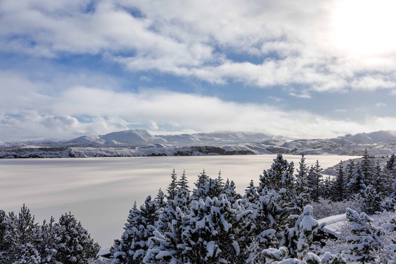 A bride and groom are walking on rocks at the base of a large tumbling waterfall. The bride is in a flowy white dress with a long veil and the groom is wearing tan pants with a red button up shirt and an orange leather jacket. The groom is holding the brides hand helping guide her along the rocks.