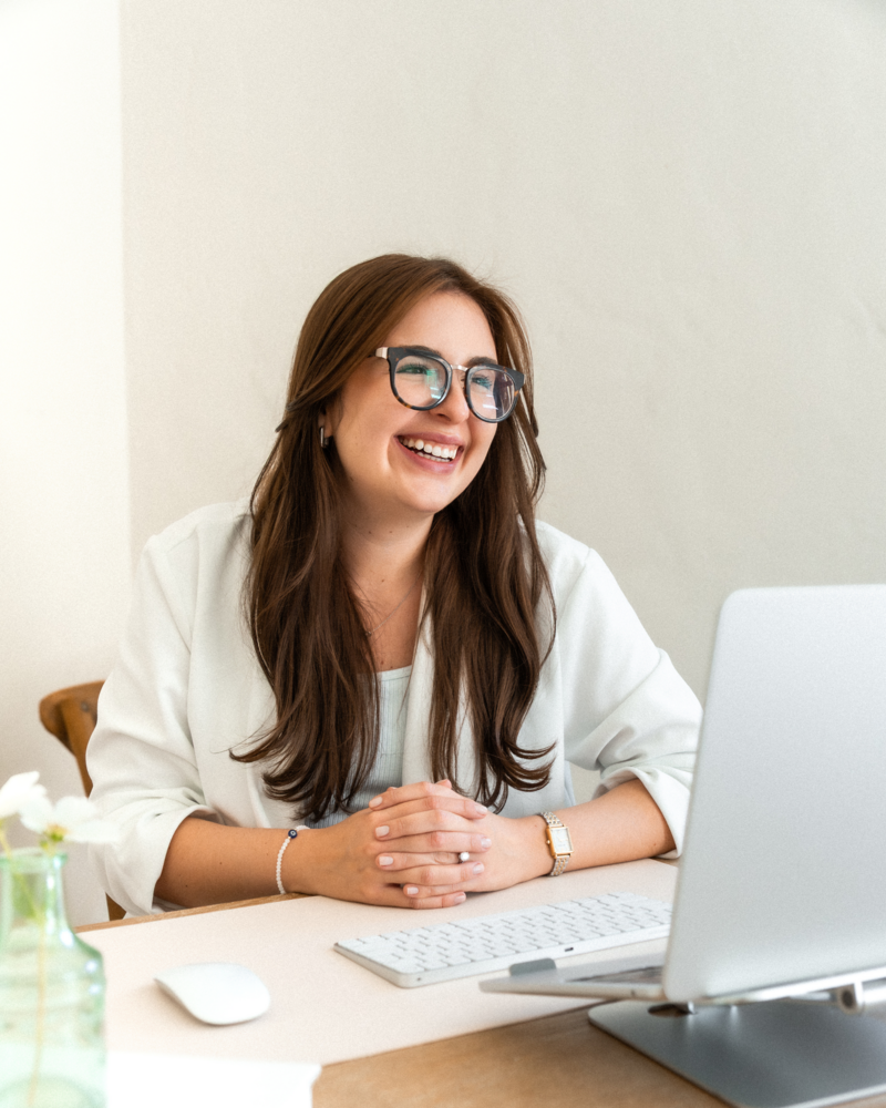 Rafa, the lead designer of Alella Creative Studio, seated at her desk with a computer monitor, keyboard, and mouse, smiling and looking to the side. A plant is on the desk.