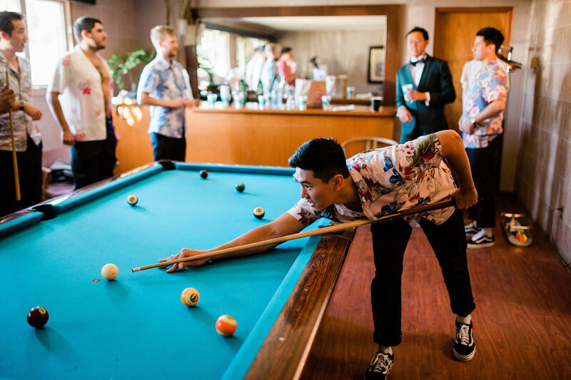 The groom and his groomsmen play pool while hanging out getting ready  in their tropical shirts and vans, with a skateboard on the floor.