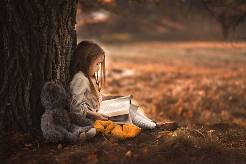 On a fall day in a park in Kamloops, a little girl sit on the ground against a tree with an open book in her lap and teddy bear by her side.  Taken by Brittany Danielle