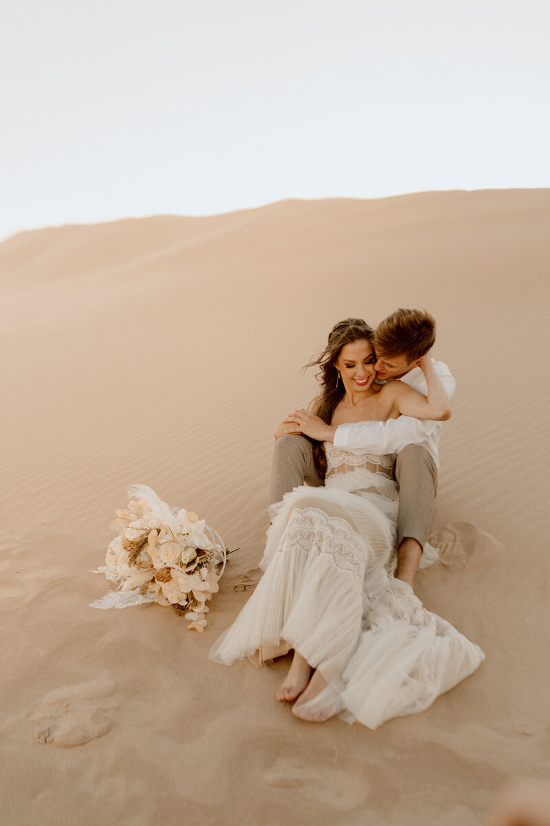 bride and groom sitting in sand dunes