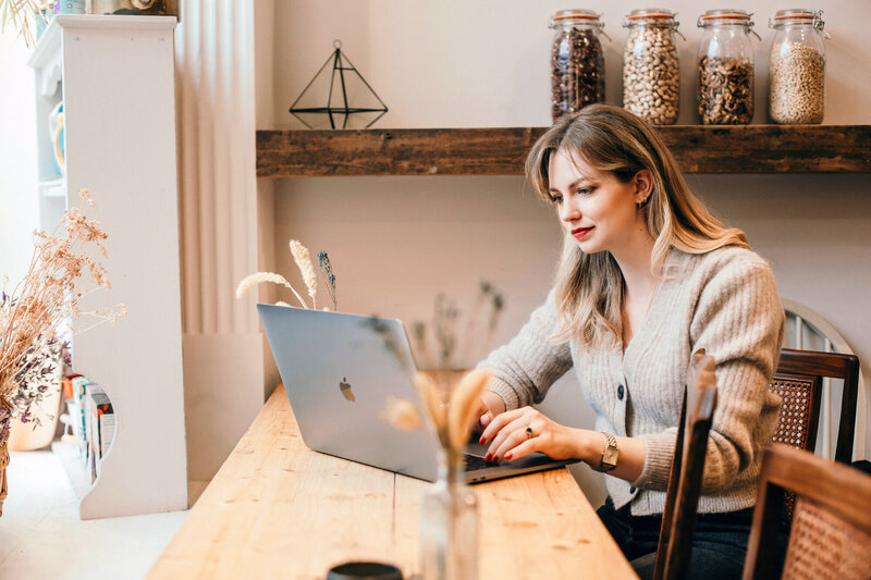  A woman's hands hold a magazine and a laptop on a wooden table, showcasing a creative workspace with a stylish aesthetic.