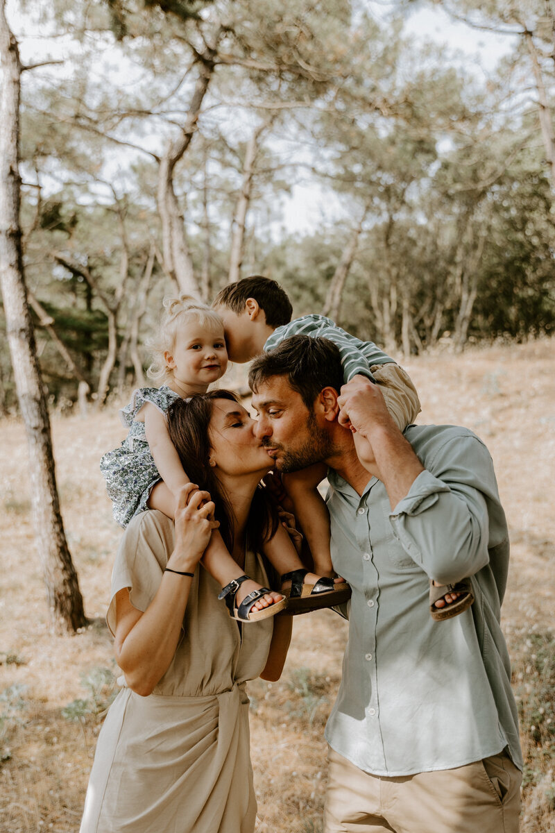 Un couple s’embrassant, portant leurs enfants sur les épaules, dans une forêt lumineuse, capturée par Laura, photographe famille en Vendée.