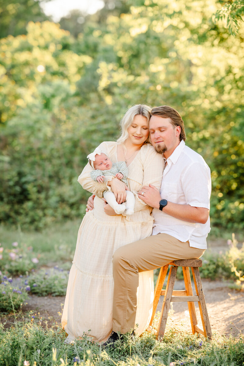 The family portrait captures a serene and intimate moment during the spring season. The scene unfolds in a field of vibrant bluebonnets, with a lush forest backdrop illuminated by the warm glow of the setting sun.  At the center of the frame, the new parents stand together, radiating joy and love. The mother, dressed in a soft, off-white dress, holds her newborn baby tenderly in her arms, gazing down at the tiny bundle with pure adoration. The father, clad in a crisp white shirt and khaki pants, sits on a stool, his arm wrapped around his partner, drawing the three of them into a tight-knit embrace.  The newborn, swaddled in a light green onesie, appears peaceful and content, their eyes closed as if lost in a peaceful slumber. The family's expressions are serene, their faces reflecting the profound happiness and wonder of this special moment.  The photographer, Bri Sullivan, has captured the scene in a light and airy style, using the natural surroundings to create a dreamlike, ethereal atmosphere. The soft, diffused lighting and the vibrant bluebonnets create a stunning contrast, adding depth and dimension to the image.  Overall, this family portrait is a beautiful and heartwarming celebration of new life, love, and the joy of family. The image invites the viewer to pause and savor the tender, intimate connection between the parents and their newborn, a moment frozen in time and preserved for generations to come.