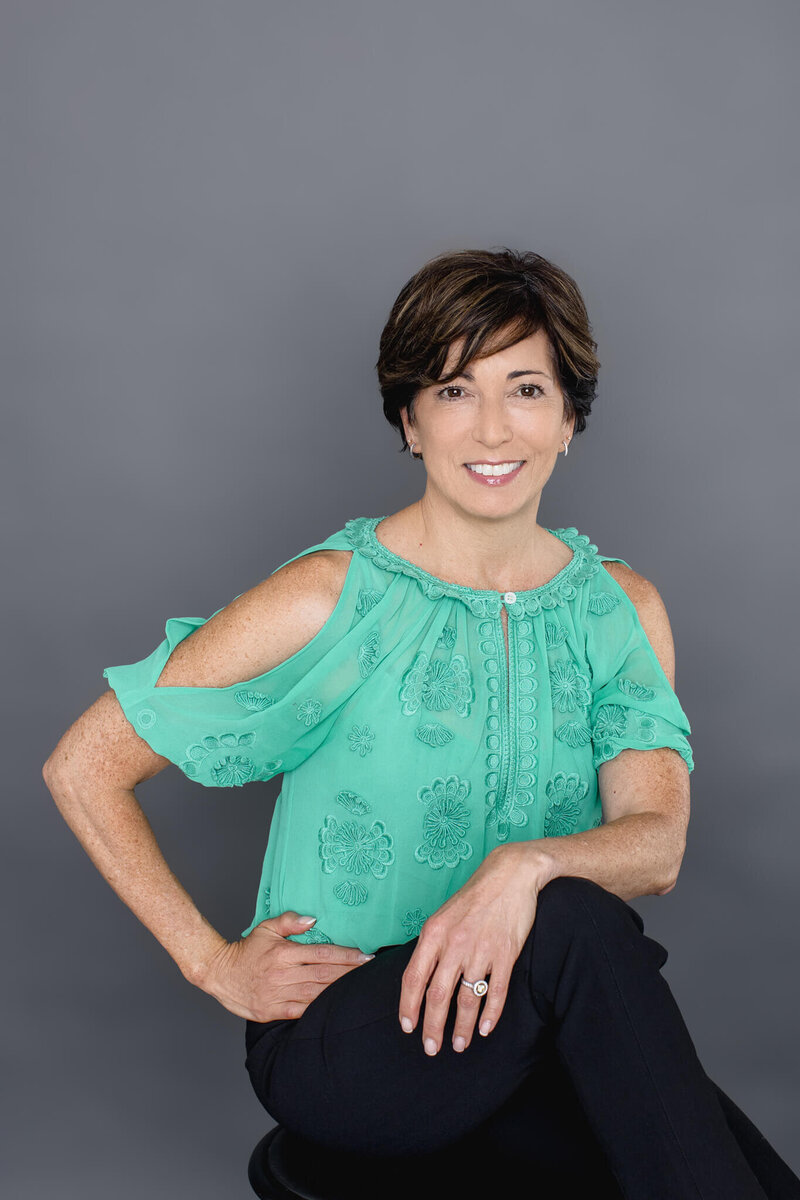 A woman with short brown hair wearing a light green blouse and black dress pants sitting on a stool in front of a dark grey background with her right arm on her hip and her left arm over her knees that are crossed, smiling at the camera.