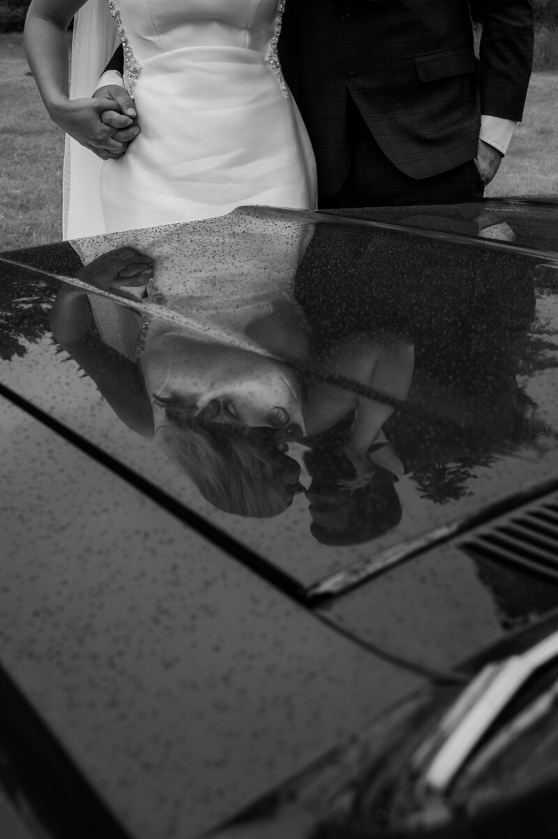 Black and white artistic reflection of a formally dressed couple on a polished car hood with raindrops, highlighting the bride’s white wedding dress and the groom’s dark suit details in a vintage-inspired wedding photography style.