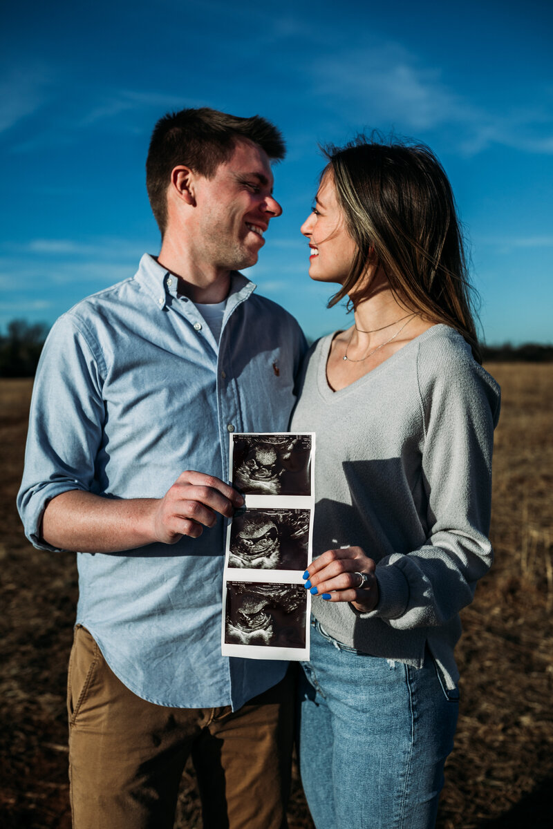 a man and woman wearing blue, outside holding a photo of their ultrasound 