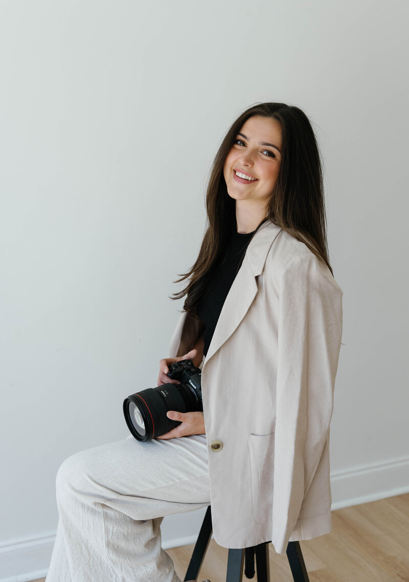 Reilly Erin, a wedding photographer based in Fort Worth, Texas, sitting on a stool with her camera in hand, smiling softly in natural light.