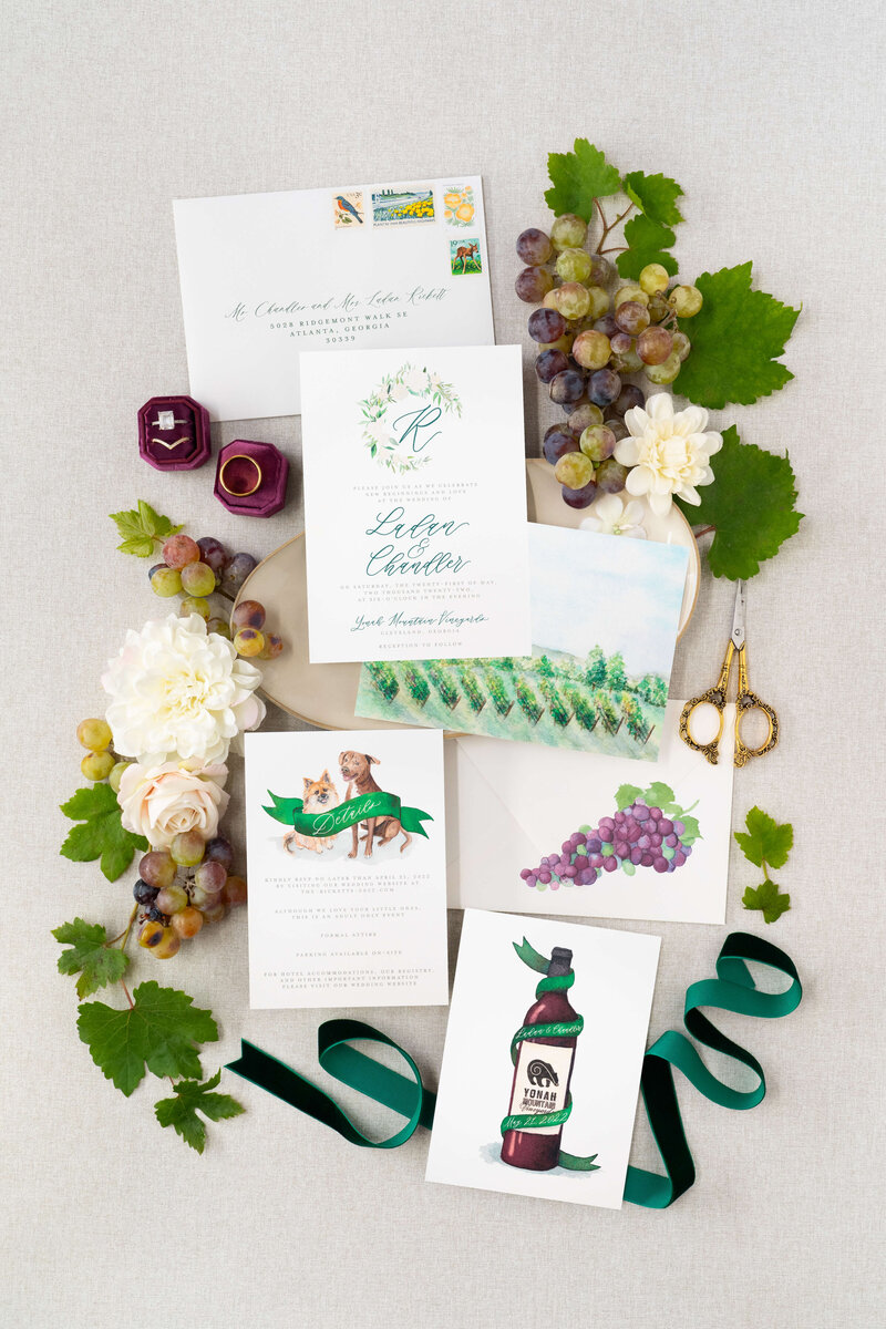 Flatlay of vineyard wedding invitations featuring custom watercolor illustrations including a vineyard scene, wine bottle, and crest, with details card showing watercolor illustrations of two dogs