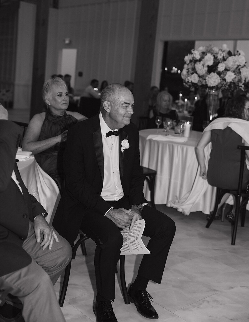 Man in a tuxedo sits smiling, holding his speech, at an Ohio wedding. Tables with floral centerpieces surround him. The atmosphere is warm and festive.
