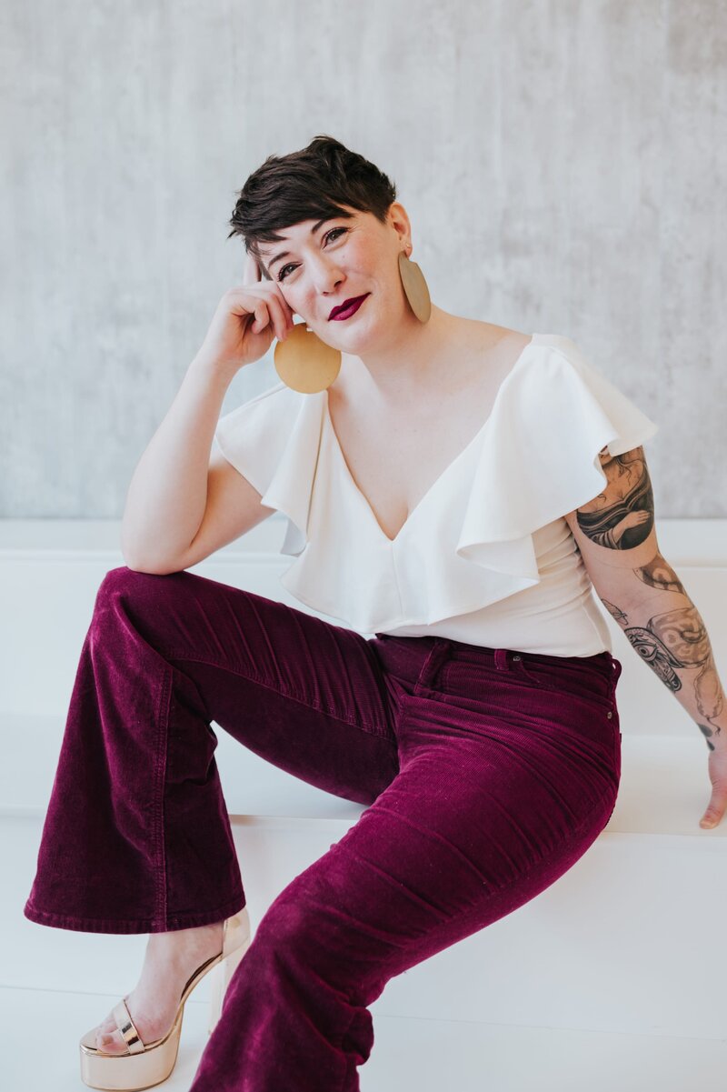 Woman smiles at the camera while wearing large, gold earrings, a white ruffled top and velvet burgundy flare pants.