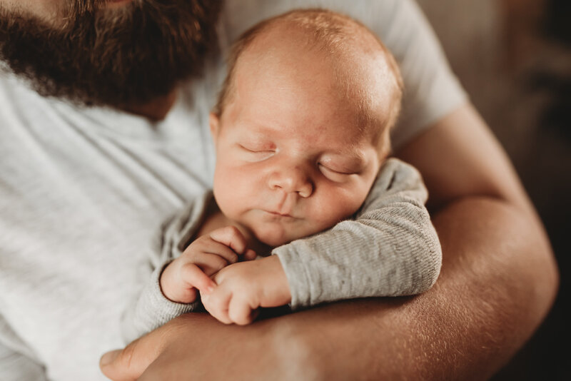 A little boy snuggles in and stays sleepy with his dads arms wrapped around him during his newborn session at the studio of nikii pix Photography. 