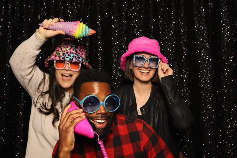 Three adults posing with colorful props and oversized sunglasses in front of a black sequin backdrop at a MISGIF photo booth during a corporate event in Phoenix, Arizona