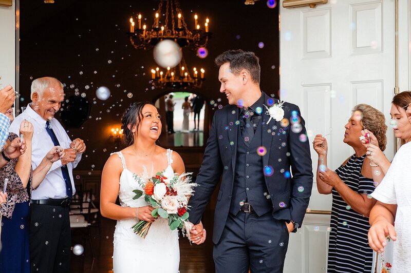 Bride and Groom laugh as they leave Maney Hall to their bubble exit from their micro wedding at Oaklands Mansion. The groom is wearing a black suit with a black shirt and white rose boutonniere. The bride is wearing a lace gown with spaghetti straps and is holding a boho bouquet of blush roses, orange roses and crimson dahlias with pampas grass and silver dollar eucalyptus.
