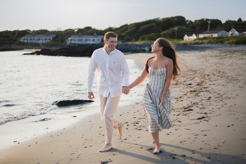 New England Wedding Photographer Pricing | Kelsey Sheehan Photography Timeless Rhode Island Weddings | A couple walks barefoot on a sandy beach, holding hands and smiling at each other. They are surrounded by calm waves and the shoreline, conveying a joyful and romantic mood.