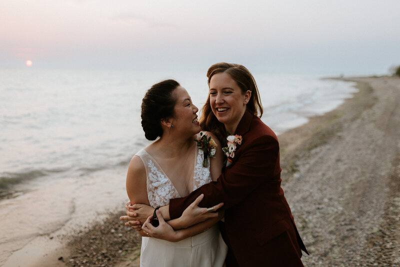 Two people, one in a white dress and the other in a dark suit, stand embracing and smiling on a beach at sunset captured by Ontario wedding photographer, Jennifer van Son at a Camp Kintail wedding.